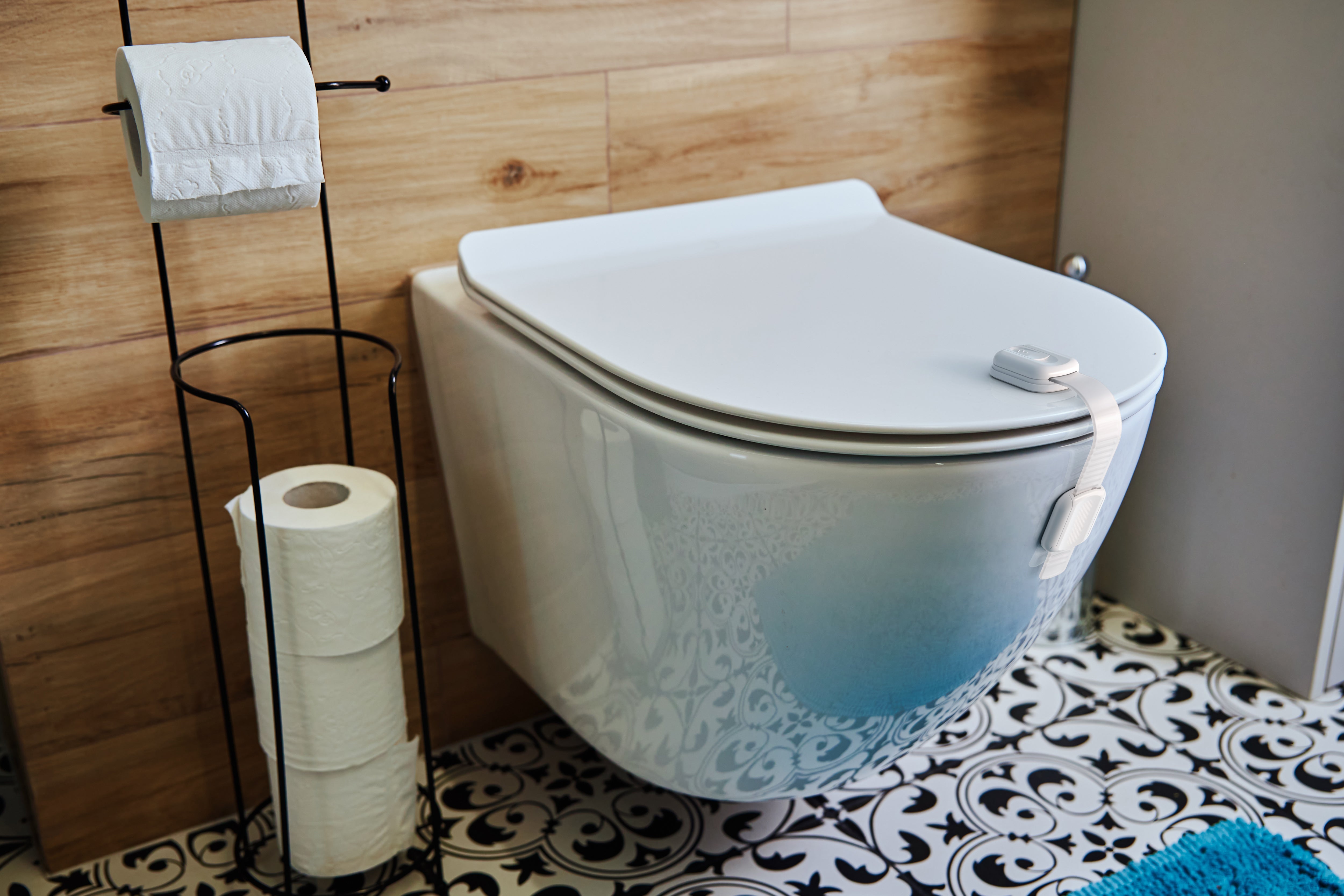 White toilet lock strap securing lid on wall-hung toilet, patterned tile floor and wood-panel bathroom walls
