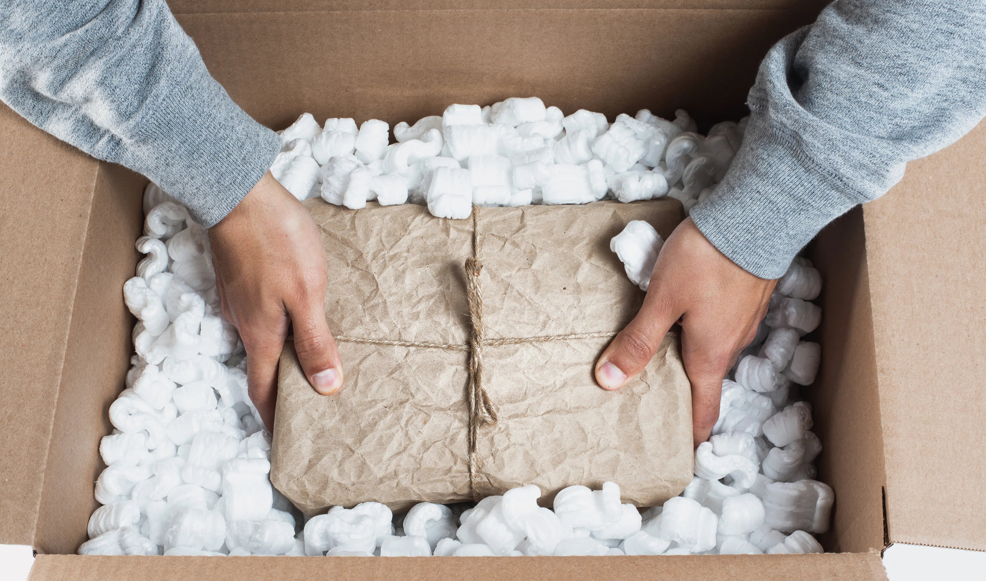 Hands lifting kraft-wrapped package from cardboard shipping box filled with foam packing peanuts